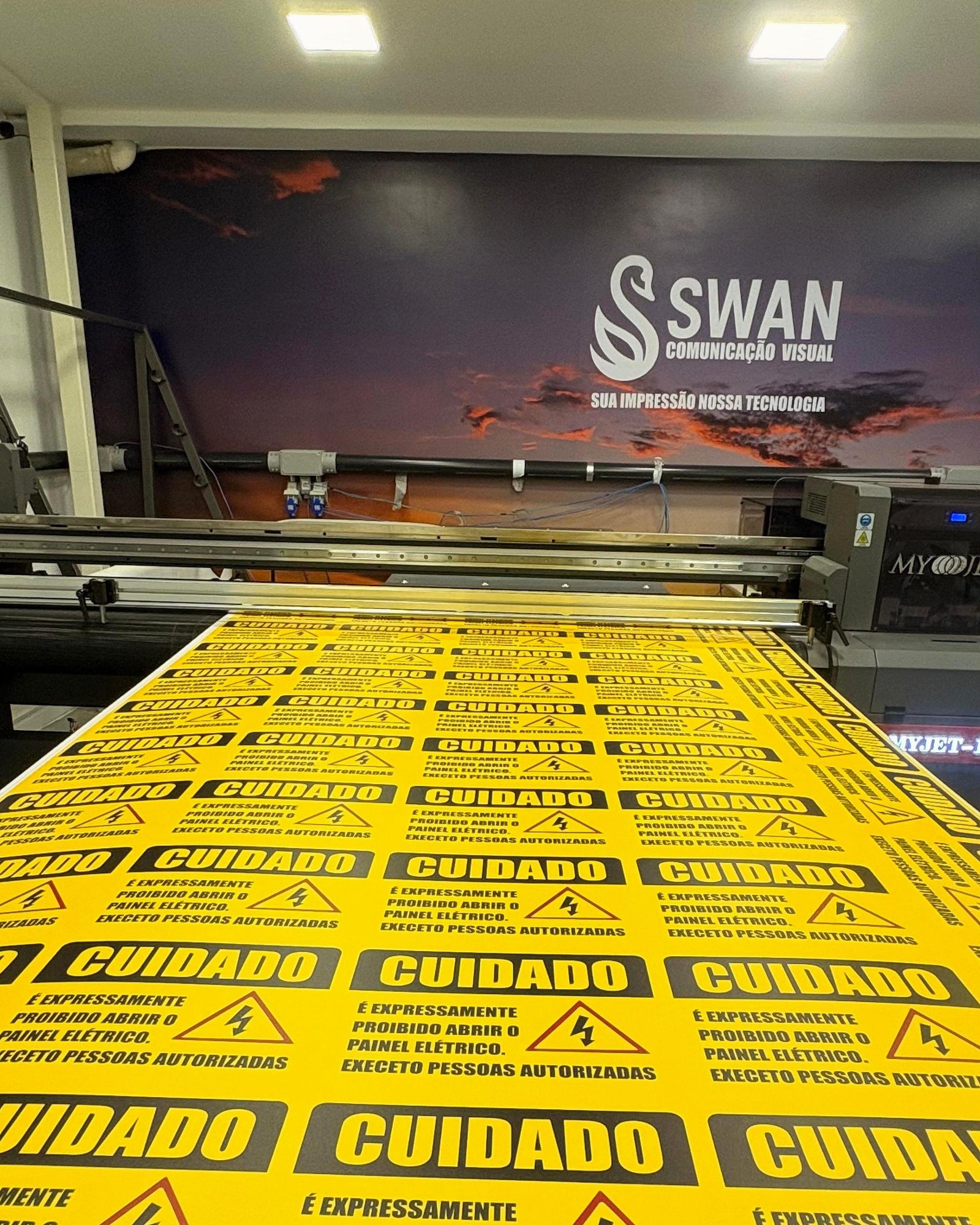 Placas de 'Cuidado' saindo da máquina, com o logo da Swan na parede ao fundo